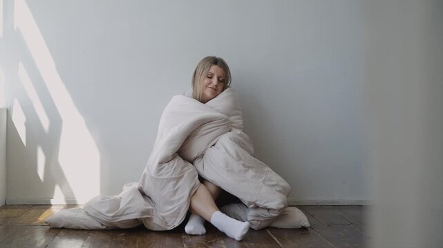 A woman sits on a wooden floor and covers herself with a warm blanket. The rays of the sun fall on the wall