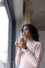 Non-binary adult standing by rainy window holding ceramic mug in pink sweater, showing smartwatch © wavebreak3