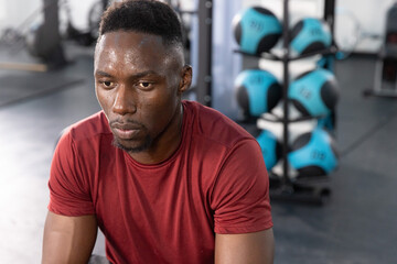 Obraz premium Adult African American man sitting at fitness center in burgundy tee, studying medicine ball rack