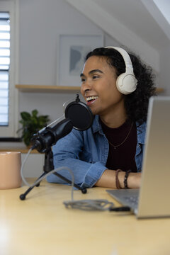 Non-binary podcaster recording at wooden desk in loft studio with microphone, headphones and laptop