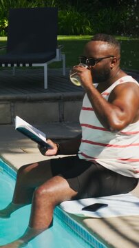 Vertical video: Reading man in striped tank sipping clear glass, glancing at open book at home pool