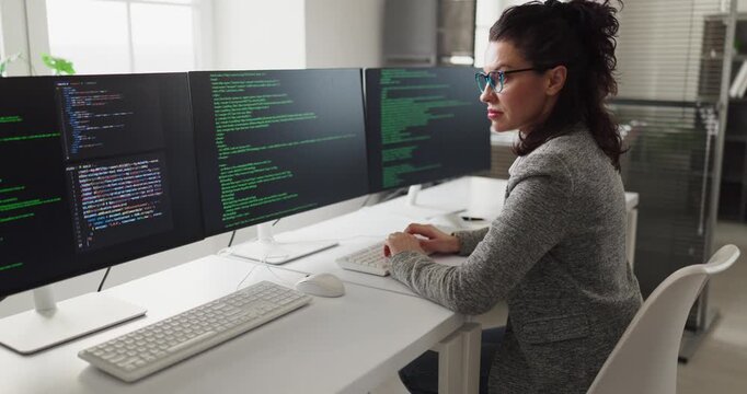Developer programmer woman coding software on screens. Side view at computer in bright office, focused coder typing at keyboard. Modern technology coding workflow concept for teams today.