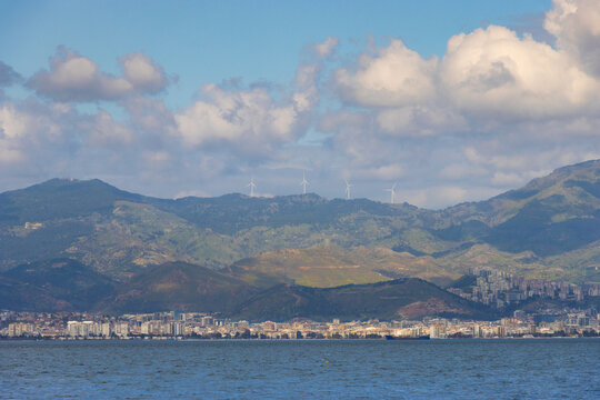 Panoramic view of Izmir city coastline with mountains and wind turbines under a cloudy blue sky across the Aegean Sea in Izmir, Turkey.