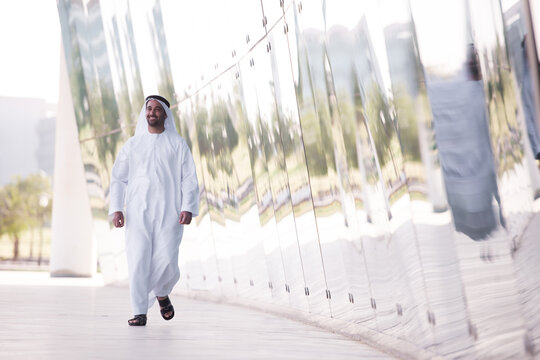 Smiling Arab man in traditional dress walking by reflective modern architecture in an urban park