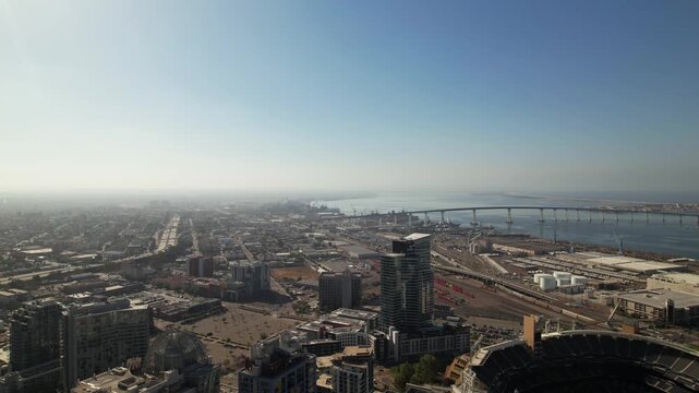 Drone flying backward above downtown San Diego at 9AM moving away from Coronado Bridge and Petco Park with wide skyline view.