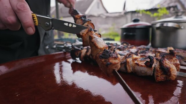 Man uses a folding pocket knife to slide a piece of succulent grilled pork from a metal skewer. The barbecue takes place in a rustic outdoor garden with pots and skewers in the background.
