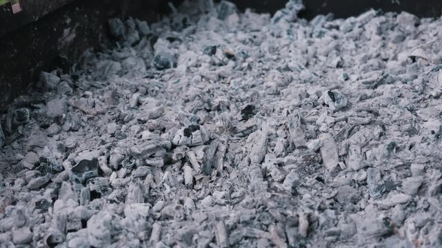 Gray wood ash and black charcoal pieces fill a metal grill, showcasing fine dusty textures and cool gray tones under soft natural light. Concept of the aftermath of fire and barbecue residue