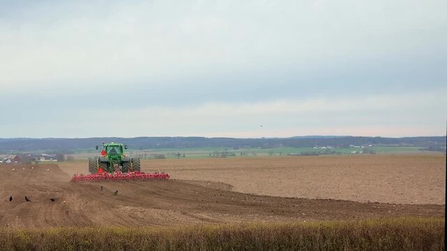 Tractor pulling a planter machine across a plowed field, seeding crops under a cloudy sky in spring