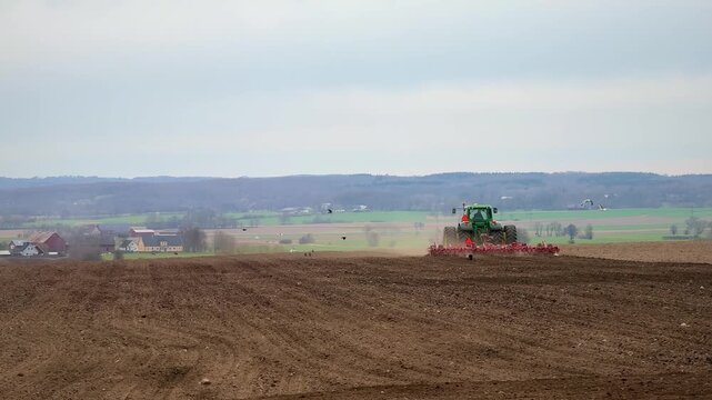 Tractor pulling a planter machine across a plowed field, seeding crops under a cloudy sky in spring