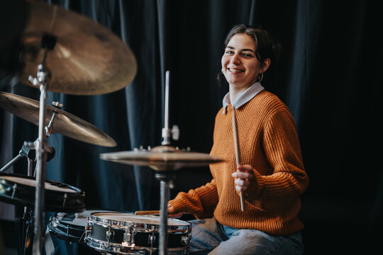 Young woman drummer playing a drum kit and smiling toward the camera. Seated at a drum set in a studio-like setting, she holds drumsticks and appears to be practicing.