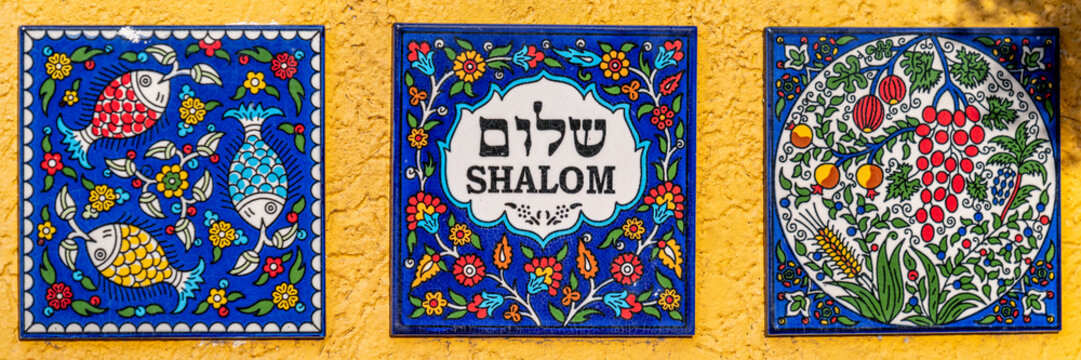 Colorful Armenian tiles one  that says Shalom in English and Hebrew text on white with a flowered blue background against a yellow wall. One tile has colorful fish and flowers on a blue background 