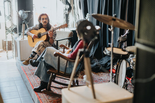 A young musician plays an acoustic guitar while a companion listens in a rehearsal space. The casual practice session shows collaboration and a relaxed, creative atmosphere.