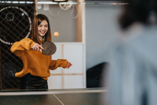 A female student serves a ping pong ball across a table while smiling. Two students play table tennis indoors in a relaxed, recreational moment.
