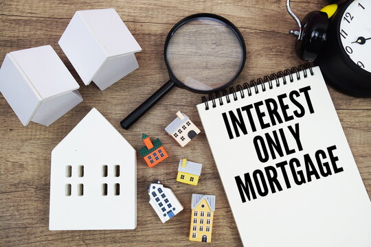 Interest-Only Mortgage text written on notepad with house model, magnifying glass and alarm clock on wooden background