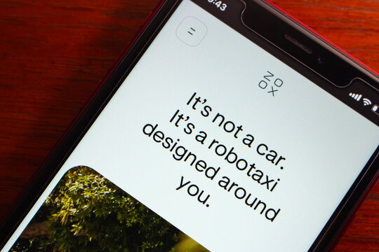 Quito, ECUADOR - Mar 2 2026 : Close-up of a smartphone displaying the Zoox website with the tagline "It's not a car. It's a robotaxi designed around you." on a red wooden surface.