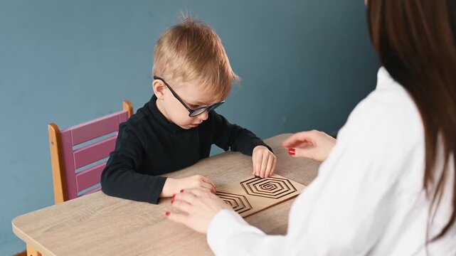 Young child wearing glasses focuses on a wooden maze activity at a table, while a teacher's hands assist in a bright classroom environment
