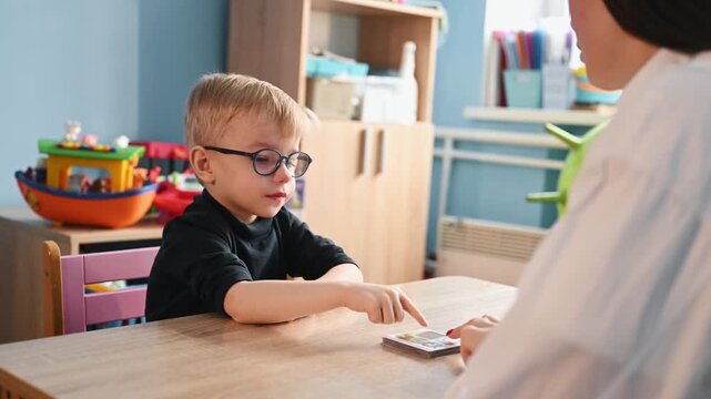 Young boy wearing glasses plays with colorful building blocks on a wooden table, while a teacher's hand assists him in a bright classroom environment