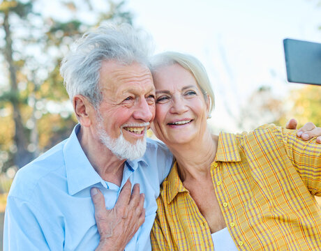 Happy active senior couple taking selfie sitting ob a bench in park outdoors. Love togetherness and acive seniors vitality concept
