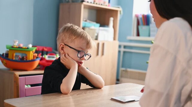 Young boy wearing glasses participates in educational activity with teacher, colorful learning cards spread on the table in a bright classroom environment