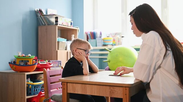 Young boy wearing glasses participates in educational activity with teacher, colorful learning cards spread on the table in a bright classroom environment
