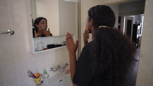 African American woman applying facial cleanser while looking into bathroom mirror during daily skincare routine creating focused self care moment in home interior