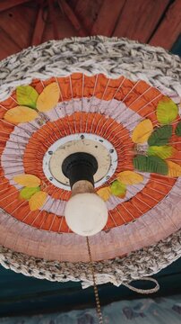 A circular pendant light fixture features a pleated paper shade with orange and pink concentric rings. Artificial yellow and green leaves decorate the surface around a central frosted light bulb