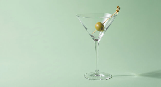 Empty martini glass with green olive on a bamboo toothpick garnish against a light green background