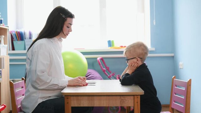 Preschool child interacts with teacher at a wooden table, focusing on educational materials in a vibrant classroom filled with toys and learning resources