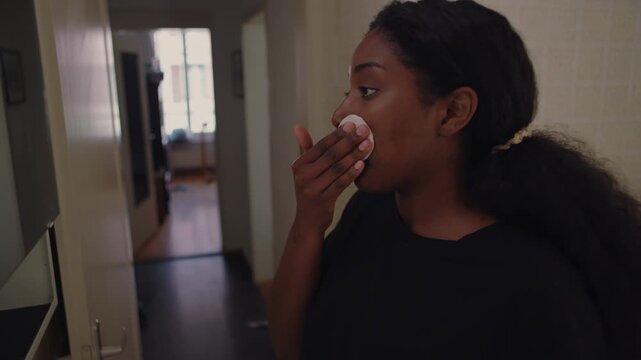 African American woman applying liquid toner with cotton pad to face after cleansing while standing in bathroom maintaining steady and mindful skincare routine at home