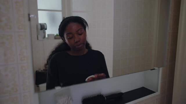 African American woman applying toner with cotton pad under eye while looking in mirror during skincare routine creating focused and delicate self care moment at home