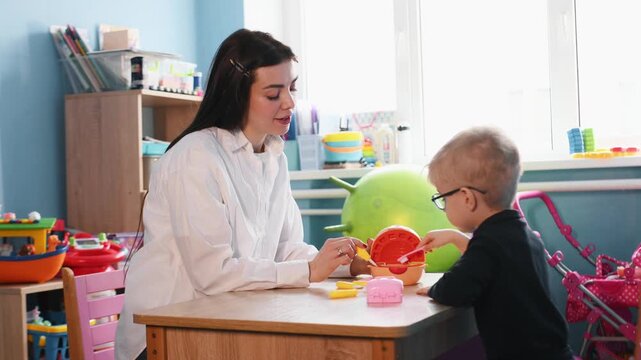Preschool child interacts with teacher during playtime at a wooden table, surrounded by colorful toys and classroom elements in a bright indoor environment