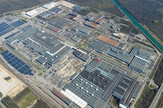 Aerial view of a sprawling industrial complex with geometric rooftops and solar panels casting shadows in the afternoon sun, Sausheim, Grand Est, France.