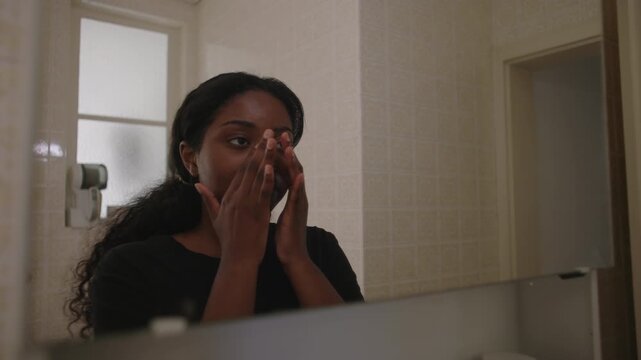 African American woman spreading facial cream across skin with both hands while looking at her reflection in bathroom mirror creating calm and attentive self care moment at home