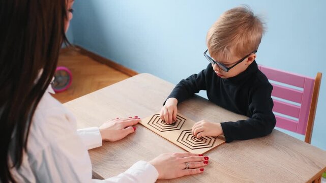 Young boy with glasses participates in a hands-on learning activity with a teacher at a wooden table in a bright classroom environment