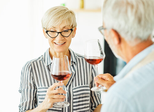 Portrait of happy senior woman and her senior edlerly husband holding wine glass and prepering meal in kitchen
