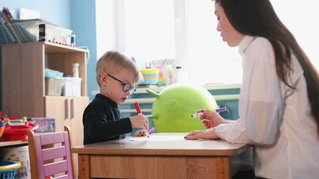 Preschool child works on a drawing with a teacher at a wooden table in a bright classroom, featuring educational toys and colorful decor