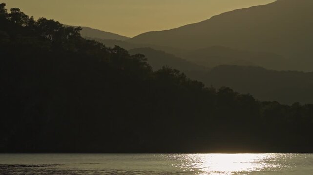 Scenic sunset over a tranquil Phaselis Bay with golden sun reflection on calm water and silhouetted hills in the background - Aerial Tele Camera View