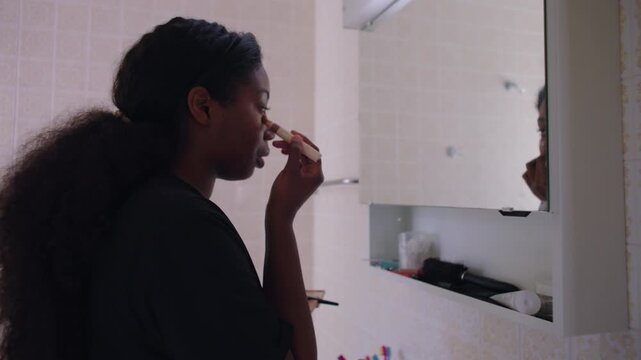 African American woman applying concealer under eye while looking at her reflection in bathroom mirror creating focused makeup routine moment in home interior