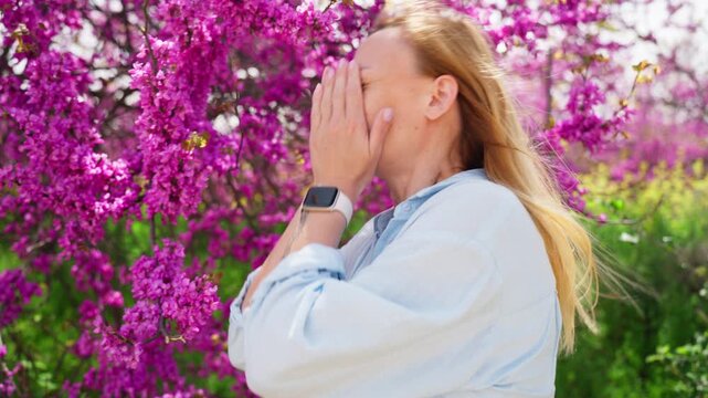 Allergic woman sneezing from pollen while smelling blooming pink flowers in spring park, experiencing hay fever symptoms, seasonal allergy reaction, and respiratory discomfort outdoors.