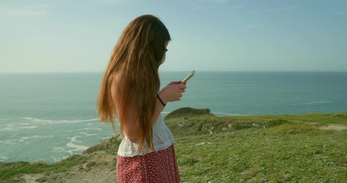 Solitude by the Sea: A young person, lost in thought, gazes at a mobile phone while perched on a cliff overlooking the endless horizon of the sea, embraced by a moment of serene contemplation.