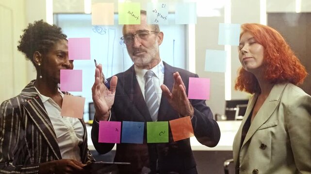 Team collaborating at an office glass wall, mapping ideas with colorful sticky notes as a leader explains strategy during a brainstorming meeting