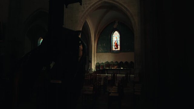 Dark interior view of the historical church in Issigeac showing rows of chairs leading to a bright stained glass window. Low-light shot.