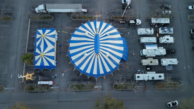 Aerial view of a vibrant blue and white circus tent pitched in a parking lot, surrounded by trucks and RVs under the soft light, St. Augustine, Florida, United States.