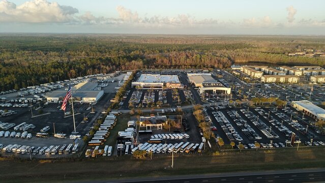 Aerial view of a sprawling RV dealership with neat rows of white vehicles contrasting against the backdrop of dense green forest under a soft, diffused sunlight, St. Augustine, Florida, United States.