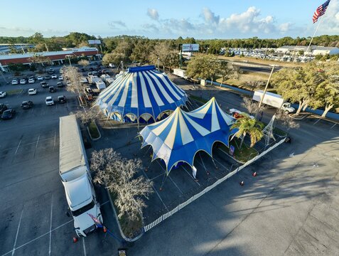 Aerial view of blue and white striped circus tents standing boldly on the asphalt, a stark contrast to the surrounding green trees and distant buildings, St. Augustine, Florida, United States.