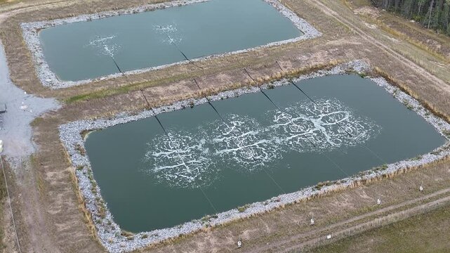 Aerial drone zooms in and orbits right around wastewater lagoons near Ilford War Lake, northern Manitoba, Canada. Aeration bubbles, berms, gravel road, and boreal forest visible.