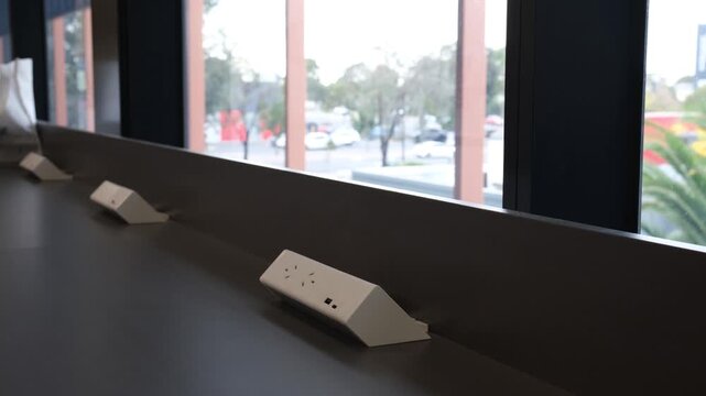 Row of power outlets installed along a desk near large windows in a modern workspace or library in Australia. Interior designed for studying or working, featuring charging points, natural light
