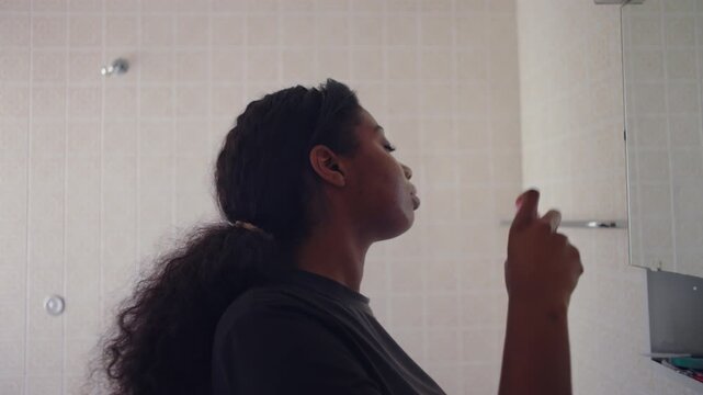 African American woman using facial spray mist while standing in bathroom with eyes closed creating light and refreshing beauty routine moment in quiet home interior