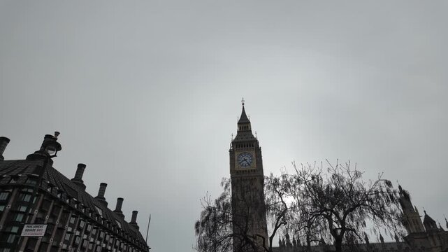 London's big ben clock tower and the palace of westminster under a cloudy sky with a bare tree silhouette, revealing the sun behind the iconic elizabeth tower landmark