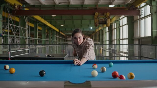 Medium shot of young Caucasian woman aiming carefully and striking cue ball at pool table in spacious industrial loft setting, showing precision and leisure activity